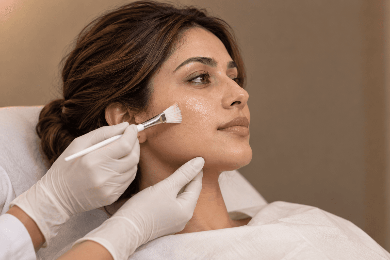 Gloved hands support a woman's jaw while a small brush applies a clear preparation to her cheek.
